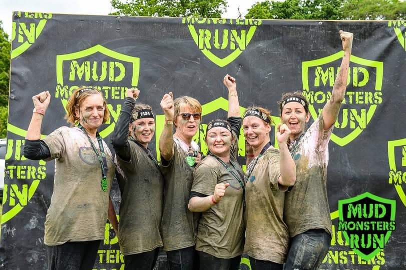 Group of women celebrating after mud run event, smiling and raising fists, with muddy clothes and a.