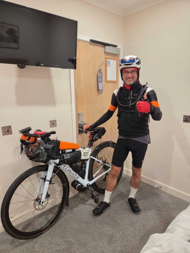 Cyclist in gear with helmet and gloves standing beside a mountain bike indoors.