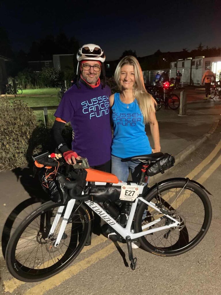 A man and woman in branded cycling gear with a mountain bike at night.